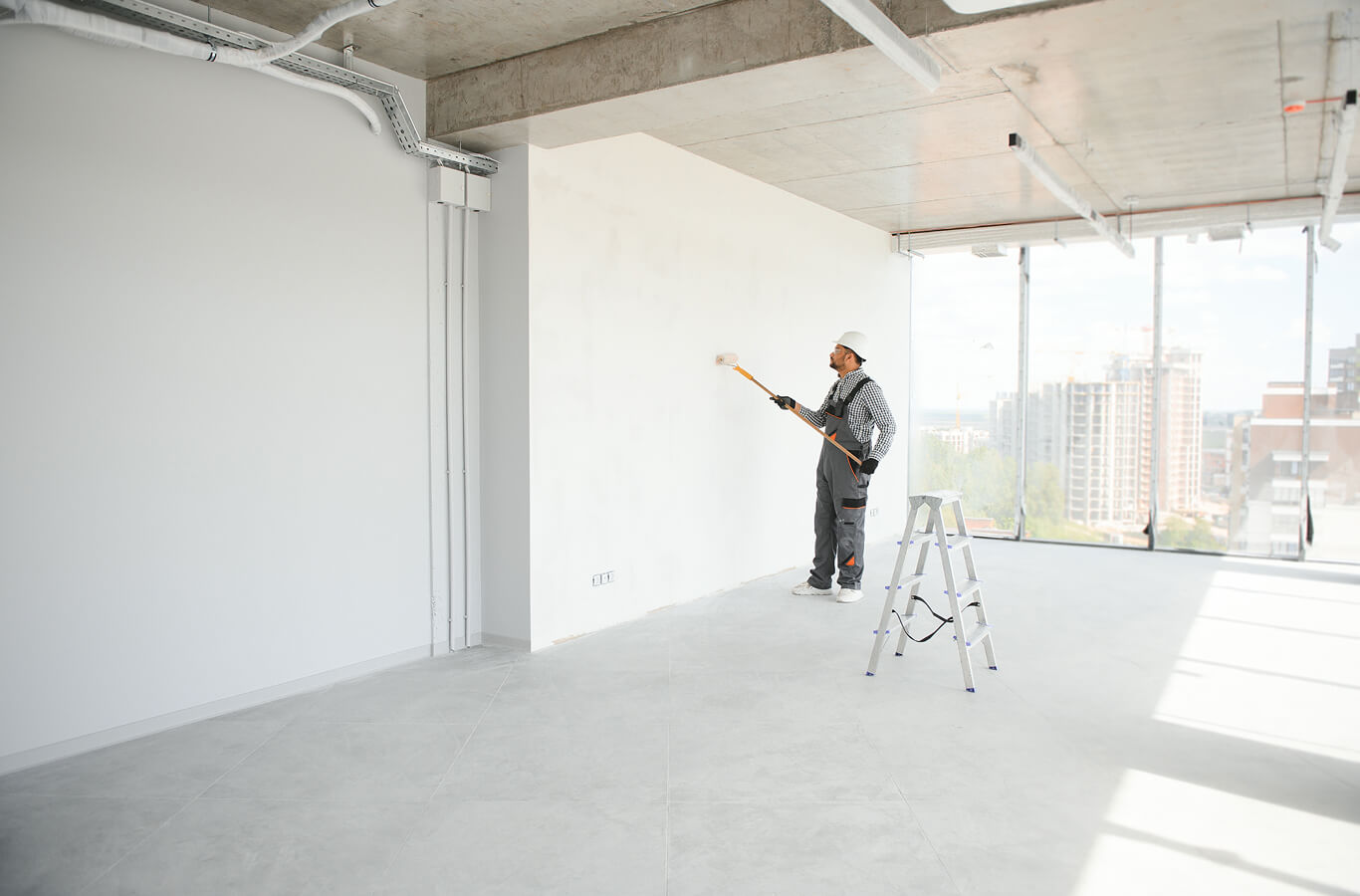 hospitality-6 A construction worker in overalls paints a white wall using a roller in a high-rise building with large windows and city views, conveying progress and modernity.
