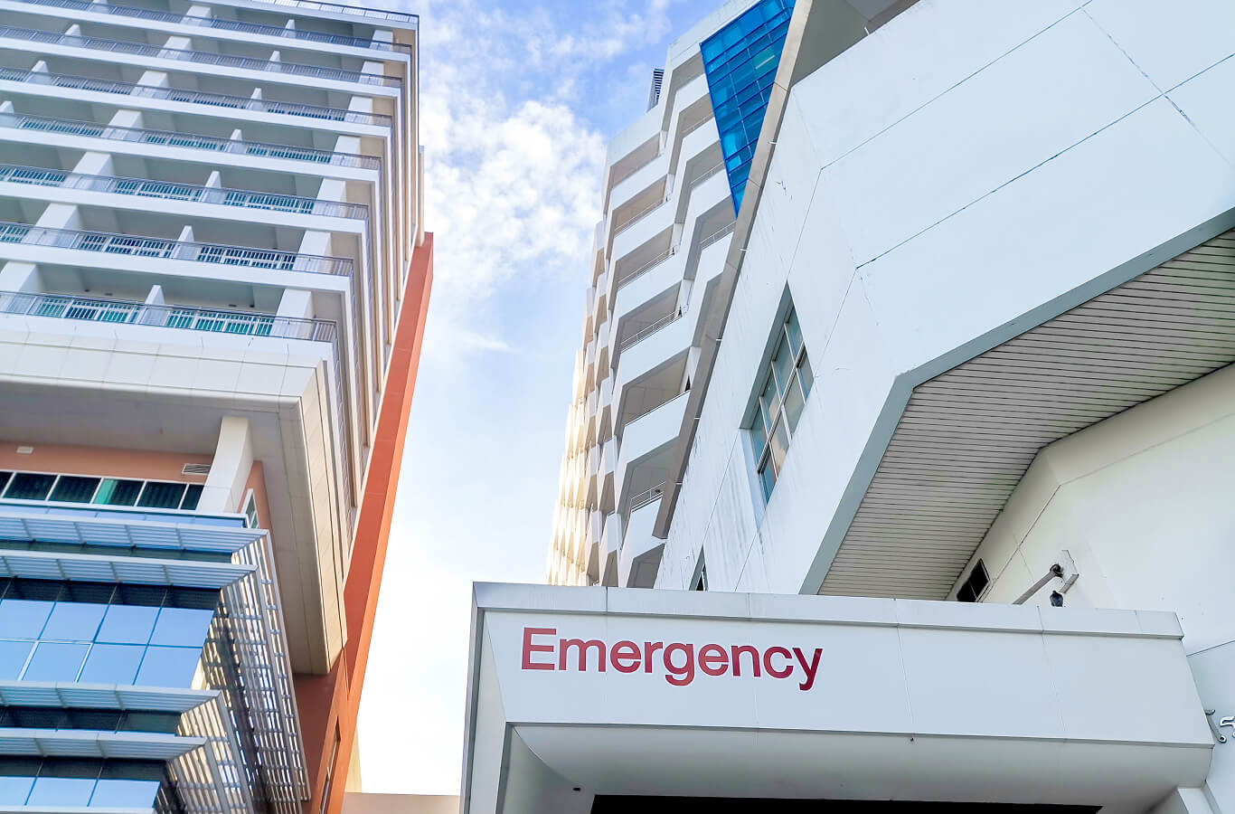 Upward view of modern hospital buildings under a blue sky, with the word "Emergency" clearly visible. The scene conveys urgency and care.