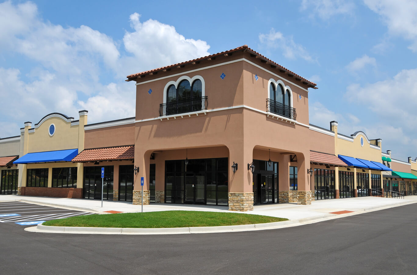 A single-story retail building with a decorative facade, featuring blue awnings and a tiled roof. The structure is located on a paved lot with marked parking spaces and green grass in front, under a partly cloudy sky.