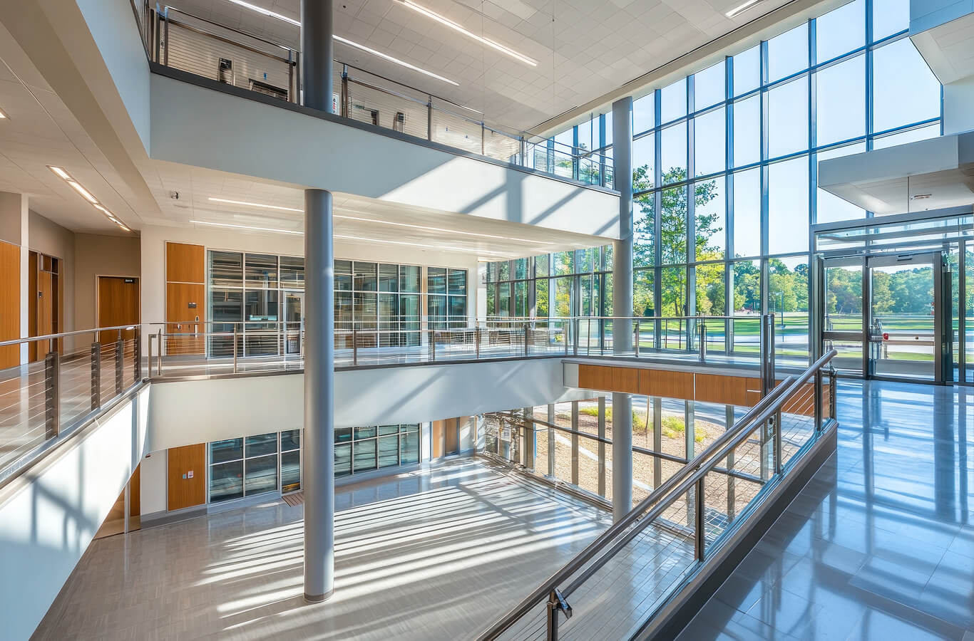Modern interior of a multi-level building with large windows, natural light streaming in, and a staircase with metal railings.